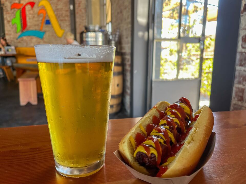 A pint glass filled with yellow beer sits on a medium-brown wooden surface next to a cooked hot dog. The hot dog is topped with yellow mustard and red ketchup and rests in a small, light brown cardboard container. In the background, there is an open glass door on the right that leads to the outside. Further back on the left, a brick wall features the letters "L.A." painted in various colors in an old English font.