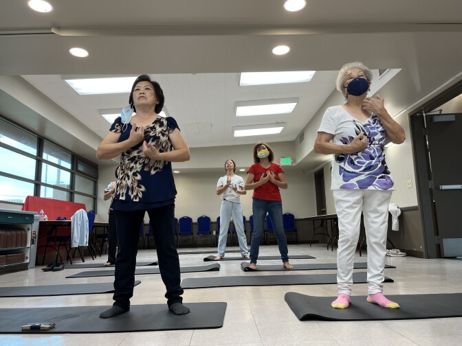 Five middle-aged Asian American women stand on black yoga mats practicing qi gong moves with their hands. 