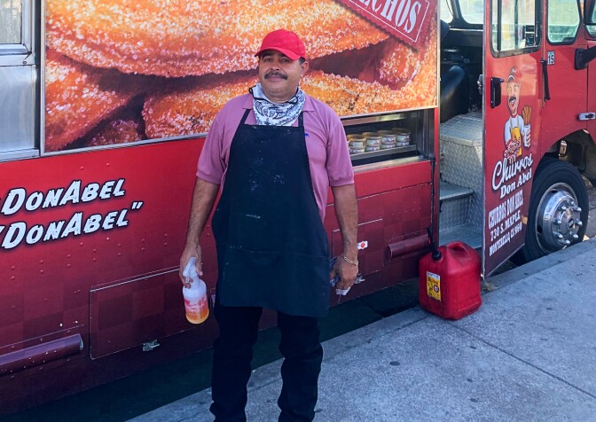 A man wearing a red baseball cap, blue apron and white bandana around his neck stands near the front of a food truck that reads "Churros" and "Recien Hechos" on the side.