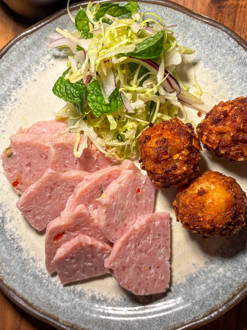 A blue and white plate displays slices of pink-colored meat, a small green salad with colorful herbs and dark golden-brown balls of fried rice.