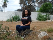 A young woman with light brown skin and curly brown shoulder-length hair wears a black t-shirt that reads "altadena" in white lettering and dark jeans. She sits on a stump in an empty dirt lot under an overcast sky. 