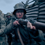 Felix Kammerer stands to the far right. He holds a rifle against his right shoulder ready to fire it if needed. Kammerer looks straightforward, eyes wide and intense, concentrating on what is in front of him. He wears a helmet and grey military uniform. His face is smudged with mud, a thin line of blood trailing along his nose. Behind Kammerer to the left is Albrecht Schuch. He too holds a rifle against his right shoulder, but its muzzle is pointed down towards the ground. His helmet hoods his eyes in shadow, and he wears the same grey military uniform as Kammerer. Behind them stands another man in the same helmet and uniform, his head and shoulders just visible between Kammerer's and Schuch's shoulders. The three men are surrounded by tall wooden and metal walls of war trenches. Everything is a grey hue. 