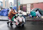 Tarps cover homeless peoples' tents on Skid Row on a cloudy day in Los Angeles. In the foreground, a woman pushes a shopping cart full of her belongings.