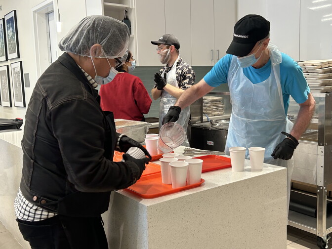Two men in masks and hairnets are standing on either side of a counter, pouring water into paper cups on orange trays. A man and a woman can be seen behind them in similar protective gear, chatting by the trays.