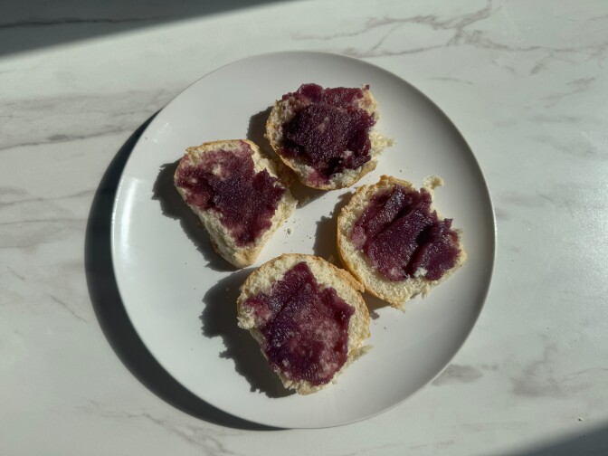 Four slices of bread spread with dark purple ube jam arranged on a white plate on marble surface with natural lighting.