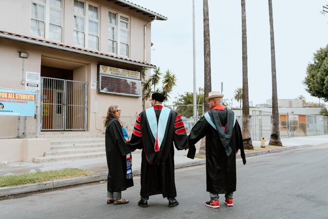 Holding hands and clad in graduation regalia, three people stand before a gated school. There are several palm trees  in the background. 