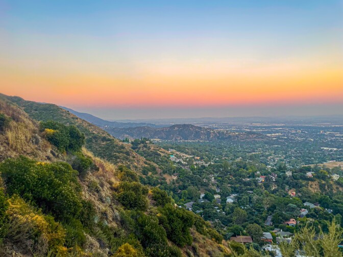 Looking towards down from the Mountain Wilson trail at dusk, a hillside green with brush sloped from the right down to the center, opening up to a view of the canyon below wich is dotted with houses and trees. Streaks of gray, pink, orange, yellow and blue line the horizon. 