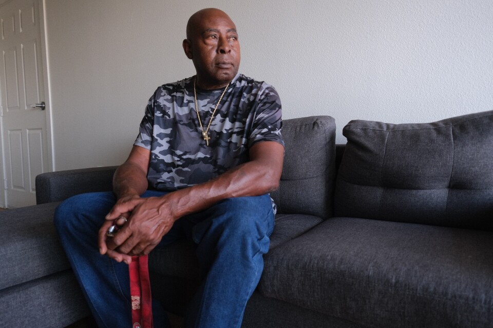 A Black man in a camo t-shirt and cross necklace sits on a dark couch with keys in his hand.