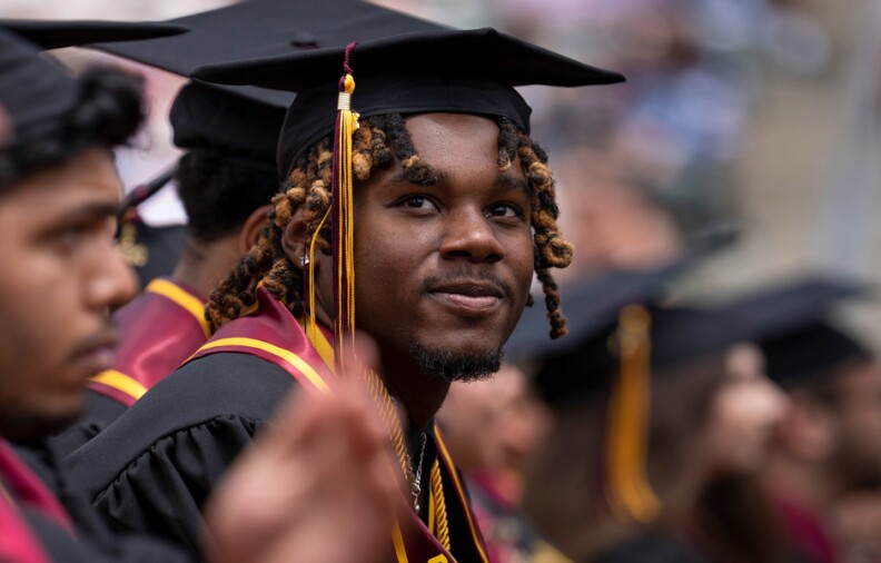 A Black student wearing graduation attire and sitting among other graduates is looking up and to the left, smiling.