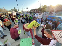 A man in the foreground holds up neon yellow signs reading 'Save Pio Pico' as dozens of people standing on a sidewalk look in his direction. Nearby, a woman speaks into a white megaphone, addressing the crowd.