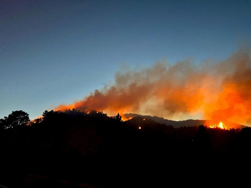 Flames and smoke from a fire are shown burning over the top of a hill in the evening.
