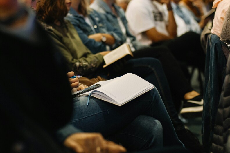 Rows of people, their faces unseen, sit and listen to a presentation. A couple o the audience members have open notebooks on their laps.