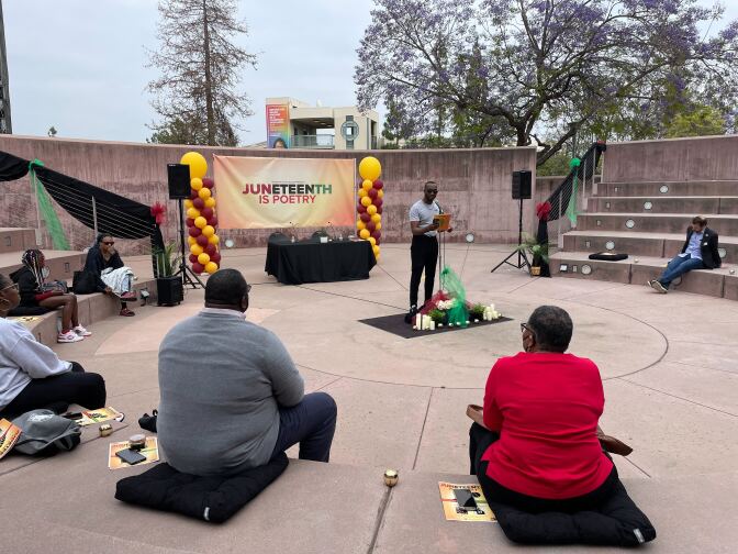 A Black man stands at a microphone in the center of an outdoor amphitheater, reading to attendees who are formed in a semi-circle.