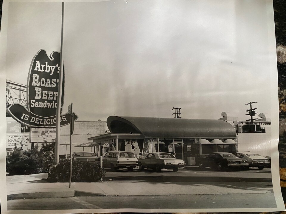 A black-and-white photo of the exterior of an Arby's fast-food restaurant, including a huge ten-gallon hat sign. At least five classic cars are parked in the lot in front of the restaurant. 