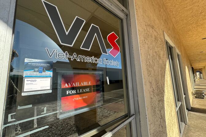 A close-up of a clear glass front door in a long, tan walkway. The door has the text "VAS Viet-America Society" displayed, with an additional sign that says "available for lease" below it.