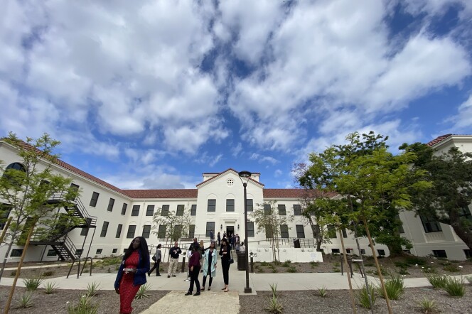 A restored historical building with two visible floors is framed by clouds above and people walking in the foreground.