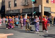 A group of women in orange curly wigs and colorful print dresses, in the style of the character Helen Roper from "Three's Company," walk down the street in a parade.