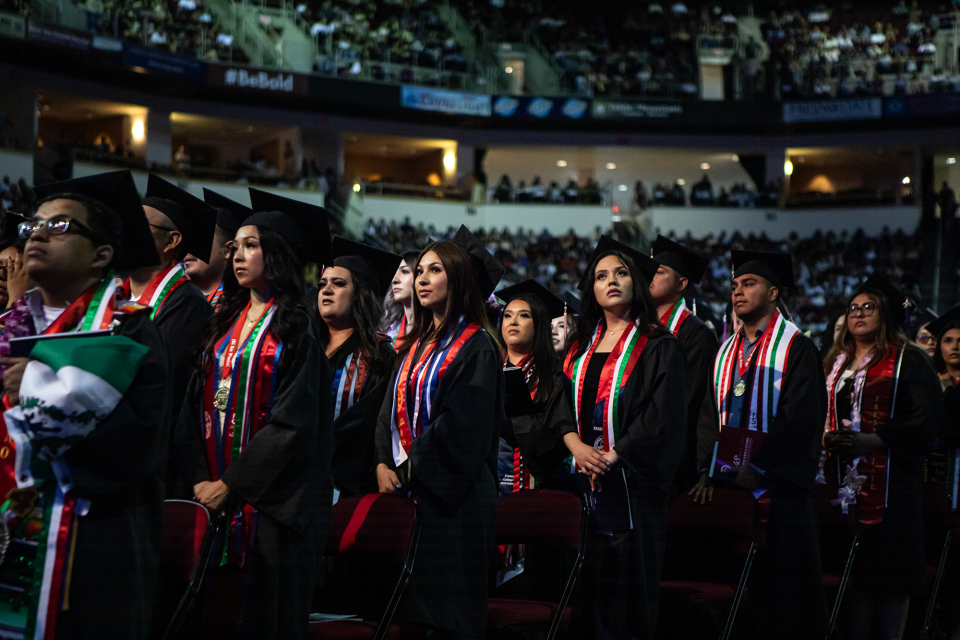 Rows of students inside an auditorium wear graduation attire.