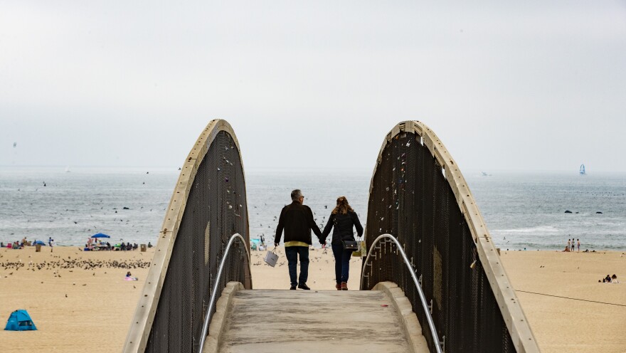 Two people hold hands while walking across a bridge. A sandy beach is in front of them with a hazy grey sky beyond.