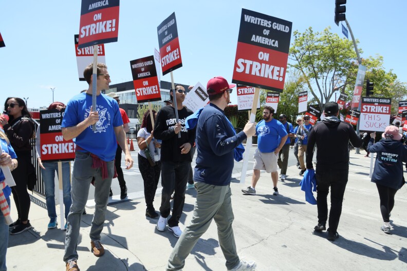 A group of men and women wearing blue T shirts and holding signs that say Writers Guild of America on strike walk around outside CBS studios, a glass fronted building in the background