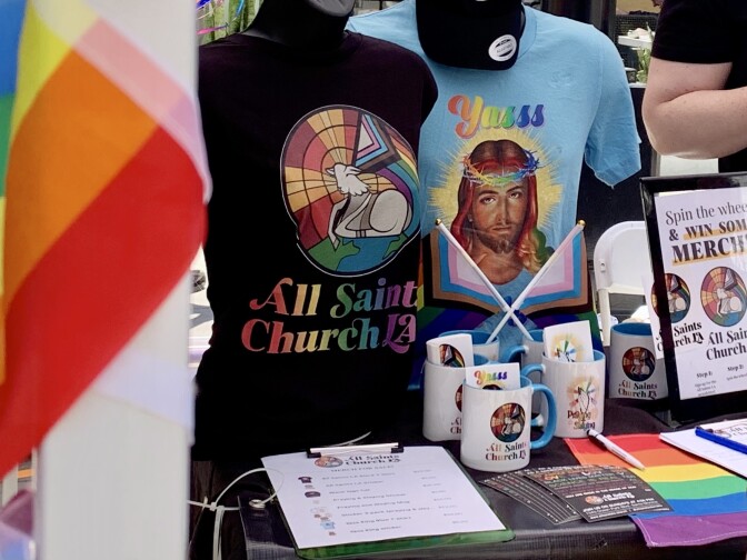 A table selling t-shirts, mugs and flags all emblazoned with various forms of the LGBTQ+ rainbow flag, along with religious imagery. One shirt features a picture of Jesus with his crown of thorns in rainbow colors and the word "Yasss" above him. Another says "All Saints Church" in rainbow lettering. 
