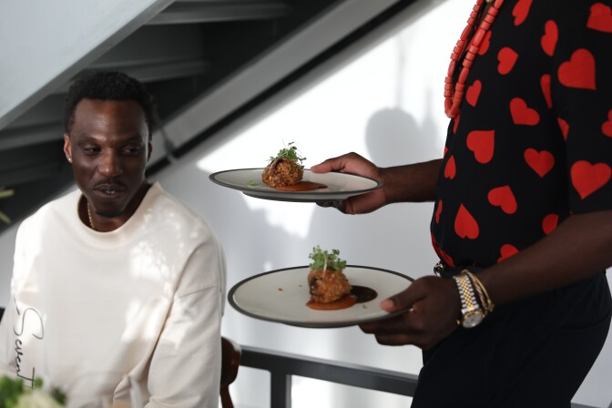 A man with dark skin is sitting wearing a white crew neck sweatshirt.  Next to him a man with dark skin wearing a black shirt with red hearts is holding two white ceramic plates, each with identical food dishes.  