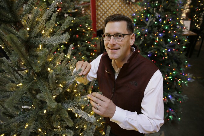 A man with light-tone skin stands next to a pre-lighted tree.