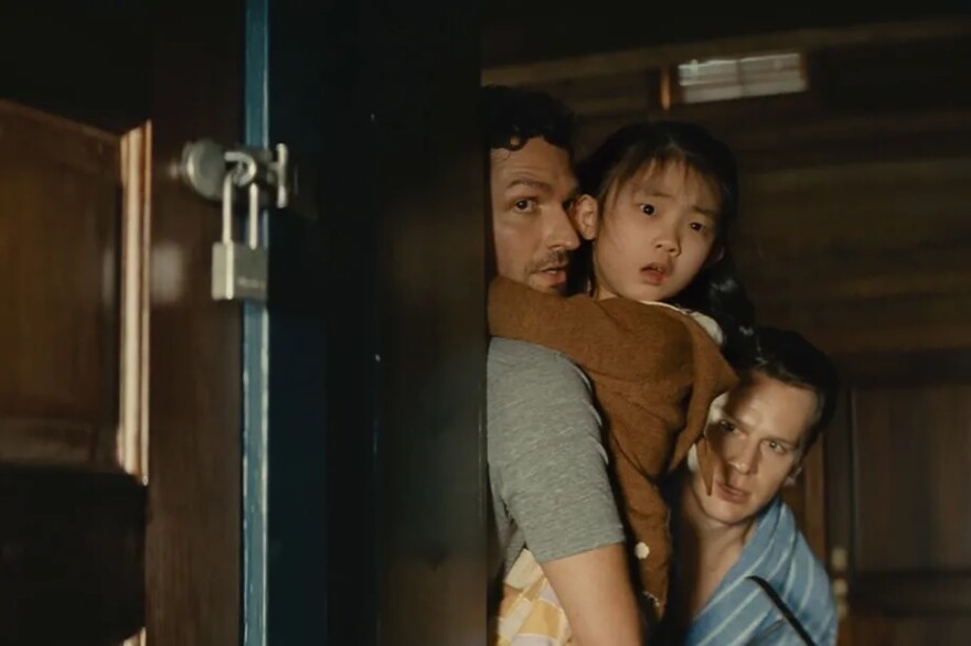 Ben Aldridge and Johnathan Groff stand in a hallway of brown wooded cabinets and doors. Aldridge stands with his back against a corner wall that leads to the back of the hallway. The wall has a door with a padlock on it to keep it shut. Aldridge wears a grey t-shirt and is holding Kristen Cui. Cui's arms are wrapped around Aldridge's neck as she looks off to the right of frame, her eyes wide and mouth open as if scared. She wears a light brown hoodie. Aldrige's face is in profile looking in the same direction as Cui, his eyes also wide with a look of concern and confusion. Groff stands slightly crouched behind the other two. He wears a light blue bath robe with white thin white stripes. He looks off to the right of frame, his eyebrows slightly furrowed with worry. He seems to be holding a crowbar. They all seem to be trying to hide behind the wall from whatever they are looking at.