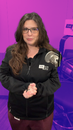 A woman with long brown hair and glasses stands in a radio studio wearing a black LAist zip-up jacket. A studio microphone and purple-tinted LAist branding are visible in the background.