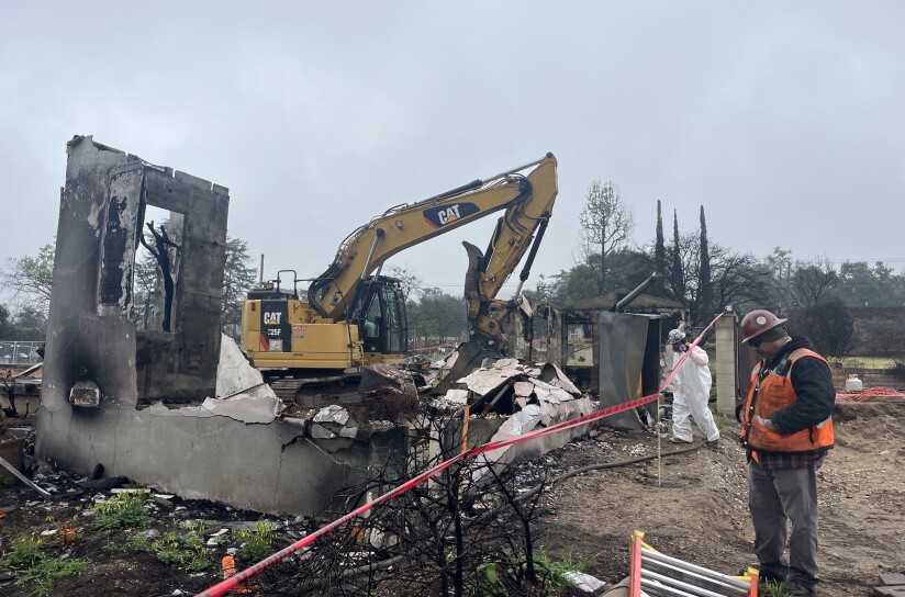 The photo depicts a construction or demolition site with A yellow CAT 325F excavator is prominently positioned in the middle of the scene. The area is filled with debris and partially demolished concrete structures.
