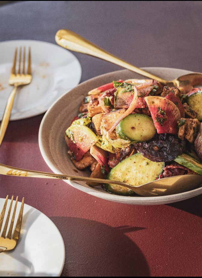 A bowl of fattoush salad with heirloom tomatoes, watermelon radish, Persian cucumber, and pita croutons, set on a dark red table with gold-handled forks and spoons nearby.