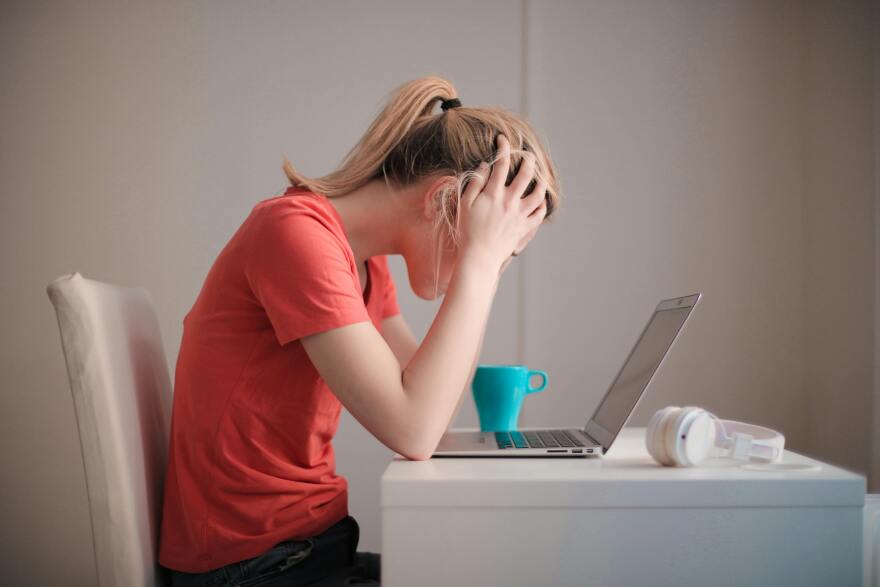 A woman in an pinkish orange t shirt sits at white desk in front of laptop holding her hands to her head in a stressed manor. A blue coffee mug and white headphones lay near on the desk.