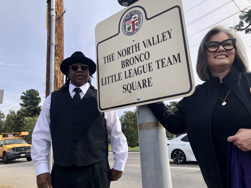 A Black man and a brown-skinned woman stand next to sign that reads "The North Valley Bronco Little League Team Square."