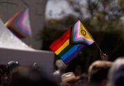 A close up of a person's hand holding a pride flag above a crowd. The flag has all the rainbow pride colors, plus these ones: A black triangle stripe for Black people. A brown triangle stripe Indigenous people. And blue, pink and white for transgender people. In the triangle's corner is a yellow area with a purple ring on it for intersex people.