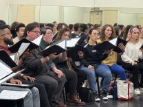 A group of more than a dozen singers with the Angeles Chorale rehearse at The Dorothy Chandler Pavilion. They hold binders filled with music notation. 