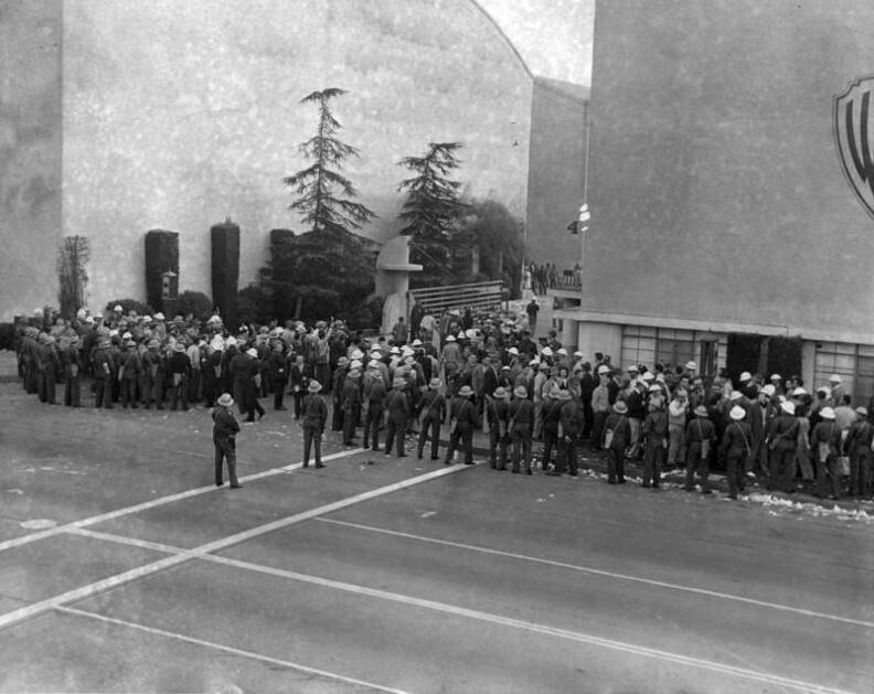 Police officers are shown standing in the street while most of the picketers are crowded on the sidewalk around the building. Police are wearing light-colored hard hats and shoulder bags, and trash can be seen strewn in the street.