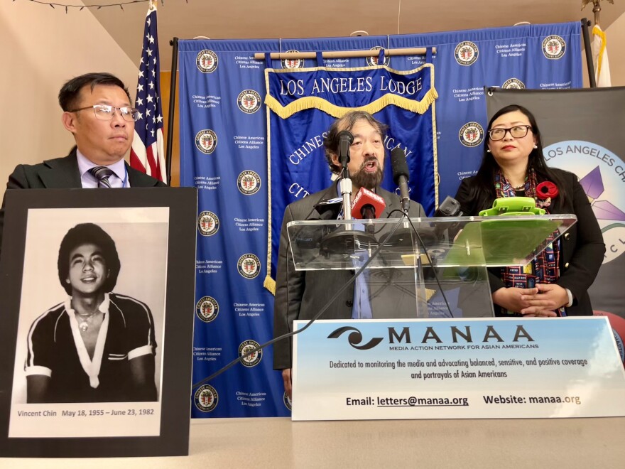 A Japanese American man speaks at a podium, flanked by a Chinese American man and a Chinese American woman. A black and white photo of an Asian American man is placed at the forefront.