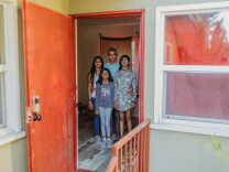 Four people stand in the doorway of an apartment with a faced dark orange door. The father, in the middle back, has his hands around the mother's waist n his right and a daughter on the left.
