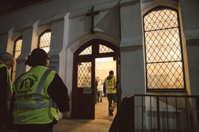 People wearing safety vests that read "Greater Los Angeles Homeless Count Volunteer" walk into a church with a white facade and a cross that hangs at the entrance.