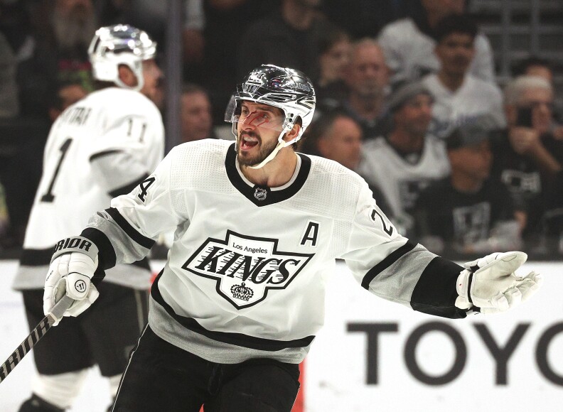 A Los Angeles Kings forward, Phillip Danault, wears a white jersey with a black border and a cross logo that reads "Los Angeles Kings" in black. He wears a chrome silver helmet with a white chin strap. He has a light brown beard and has a cut on his face which is dripping blood.