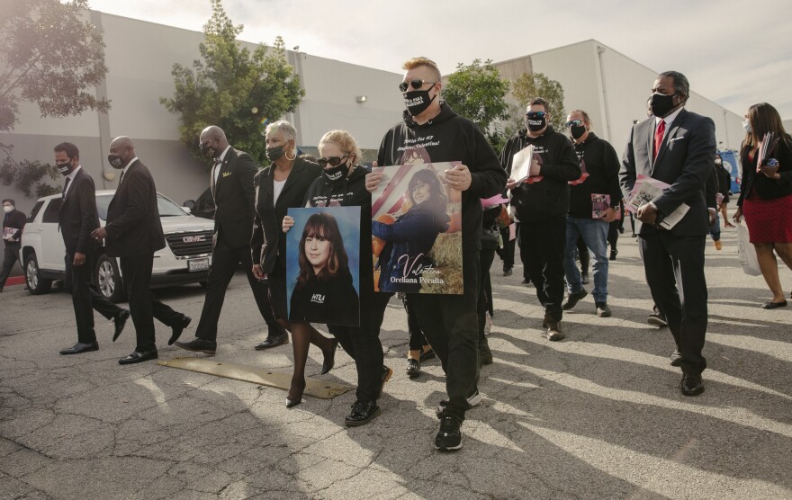A small group of people, all dressed in black walk together in what looks like a parking lot. A man and a woman at the head of the group carry large printed photos of a young girl. One photo reads "Valentina Orellana Peralta."