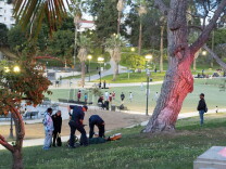 Two paramedics treat a man laying down in the grass. Boys play soccer in the background.