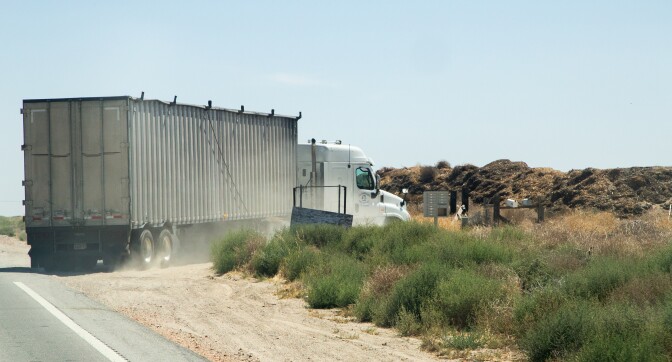 A large white semi-truck turns into a property surrounded by short dry bushes and large piles of dirt.