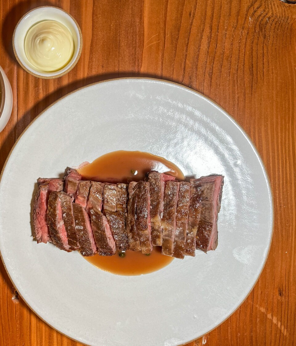 A collection of plates is arranged on a medium dark brown surface. In the upper right corner, there are golden-yellow French fries accompanied by a small container of white sauce that resembles mayonnaise. In the center, a large round white ceramic plate holds slices of cooked brown steak, drizzled with a light brown sauce.