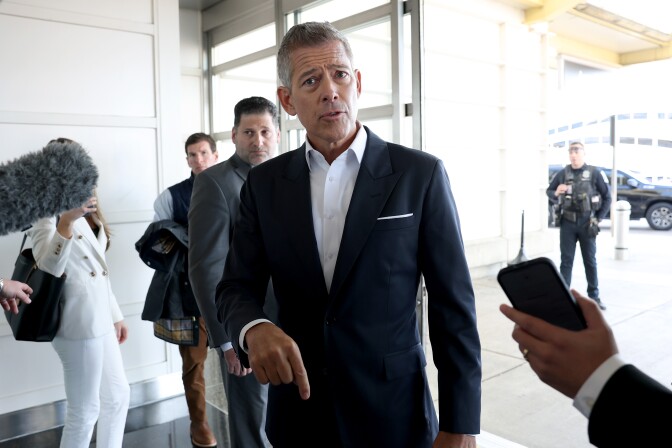 A man wearing a dark suit stands speaking with reporters holding out a cellphone and a grey, fuzzy microphone.