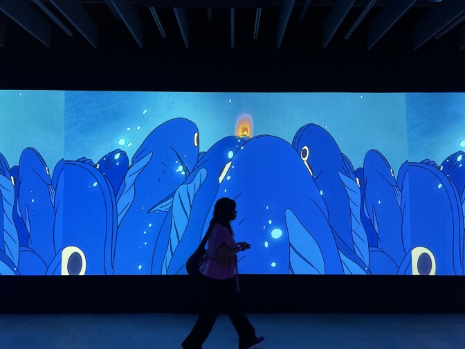 A large screen that takes up the entire wall is showing animated blue fish swimming up towards the top of the screen, with a small red and orange character just above the fish. A woman wearing a bag over her shoulder is walking in front of the screen.