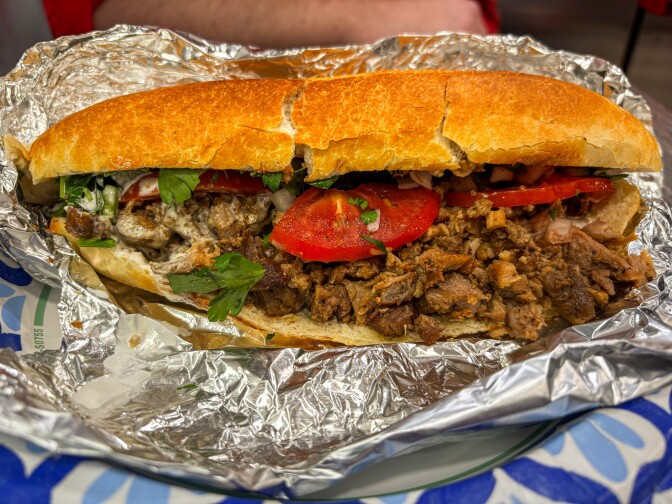 A large sandwich on toasted bread filled with seasoned beef, fresh tomato slices, parsley, and onions, wrapped in foil on a plate.