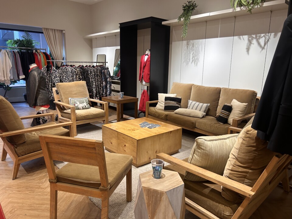 The interior of what appears to be a suit shop, with two tan couches and three chairs set up around a light brown square coffee table. There are racks of suit jackets hanging in the corner, near the front window.