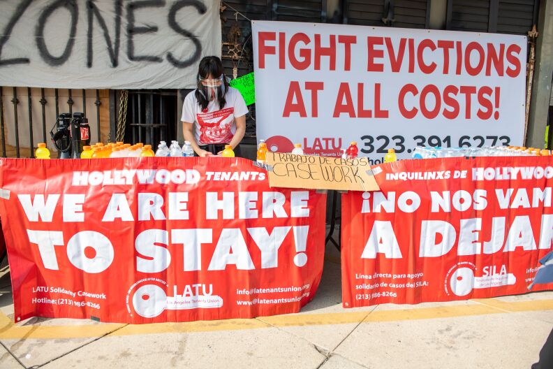 A person in a plastic face shield stands in the middle of big banners in red and white that read: "Hollywood tenants we are here to stay" and "fight evictions at all costs"