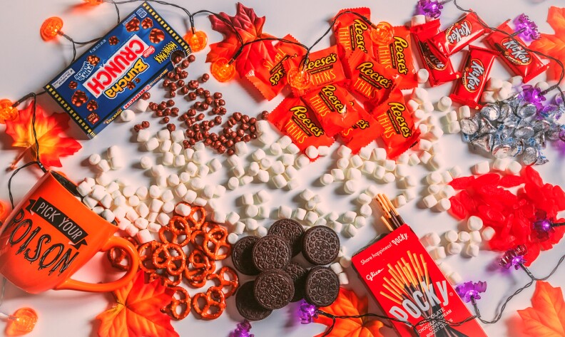 An array of candy is spread out on a table decorated with Halloween decor. Candy includes Crunch, Pocky, Reese's, KitKats and Oreos, as well as pretzels and marshmallows.